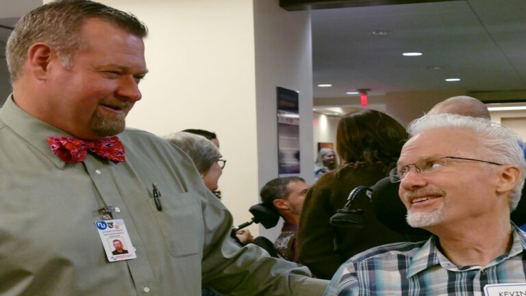 A man in a green shirt and red bow tie smiles while talking to a man with white hair and glasses in a plaid shirt, who is seated in a wheelchair. Stuarts Story unfolds as other people are visible indoors in the background.