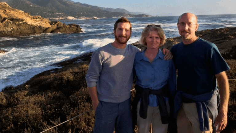 Three people stand together smiling in front of a rocky coastline with ocean waves and distant hills under a clear sky, capturing a moment from Chucks Story.