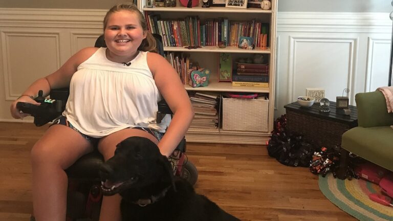 A smiling girl sits in a wheelchair indoors with a black dog by her side, bookshelves and decorative items behind her on wooden flooring, sharing a moment from Carlees Story.