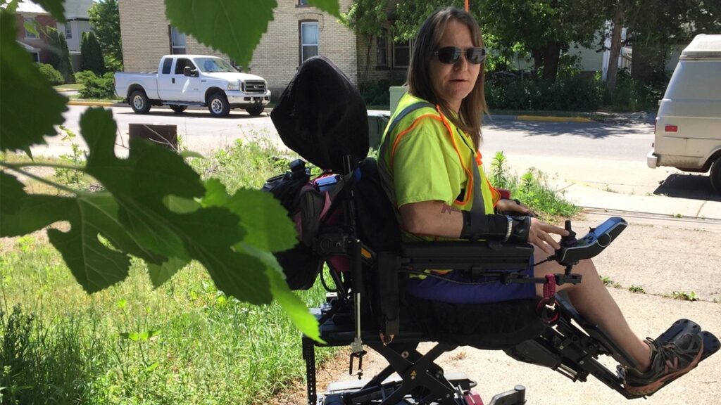 A woman wearing a neon safety vest and sunglasses sits in a motorized wheelchair on a sidewalk, holding the controls. Green leaves partially frame the foreground, and vehicles and buildings are visible behind her—this is Briannas Story.