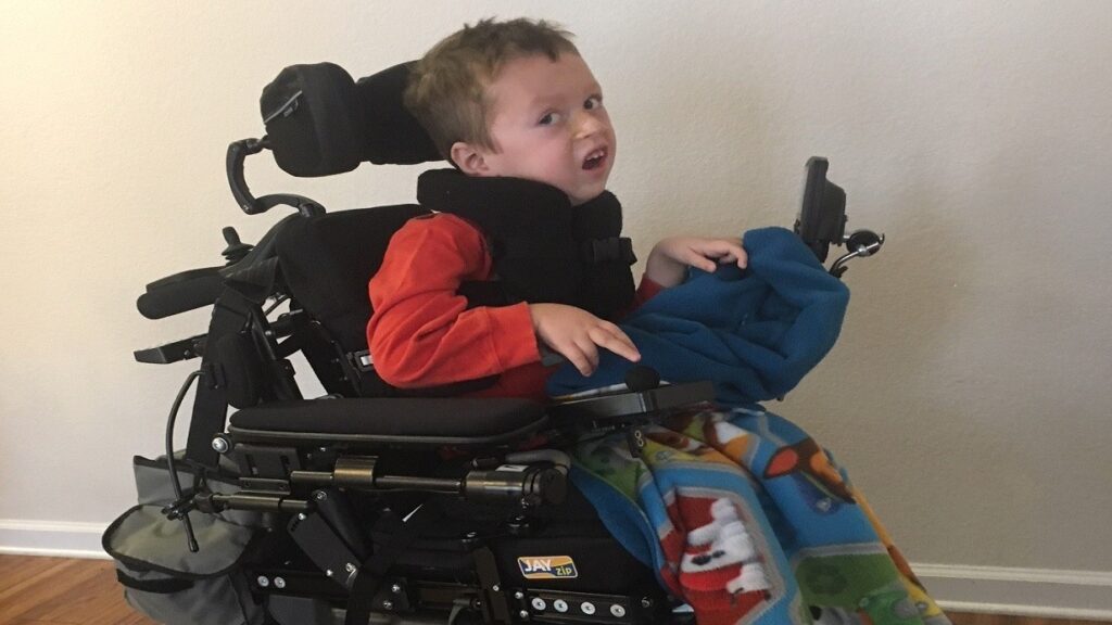 A young boy in a motorized wheelchair wears a red shirt and holds a blue blanket. He has light brown hair and sits near a white wall, with a colorful blanket over his legs, capturing a moment from Brogans Story.