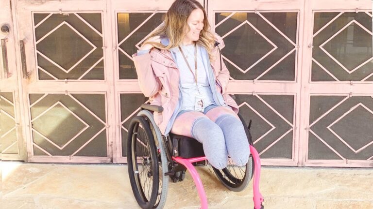 Callies Story: A woman with prosthetic legs sits smiling in a wheelchair with pink accents, wearing a light jacket and gray pants, in front of a decorative pink metal door.