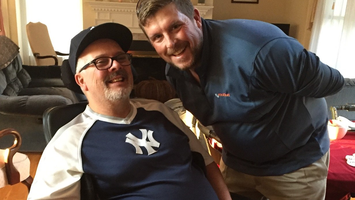 Two men smiling indoors; one is seated and wearing a Yankees shirt and cap, while the other leans in beside him in a blue shirt. The background reveals a cozy living room with furniture and a fireplace, capturing a warm moment from Careys Story.