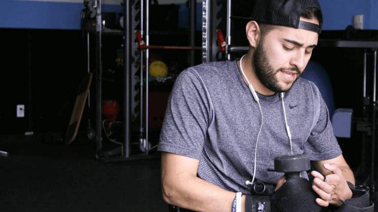 Carloss Story: A man in a wheelchair, wearing a backward cap and workout clothes, lifts a dumbbell in a gym with equipment visible in the background.