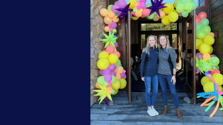 Two women stand smiling with arms around each other in front of a doorway decorated with colorful star and flower balloons, celebrating Colleens Story. The left half of the image is a solid dark blue color.