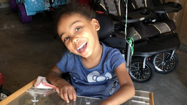 Young girl with short curly hair and a big smile sits in a wheelchair, wearing a blue shirt with a white heart. She is indoors, with strollers and toys visible in the background—part of Kianas Story.