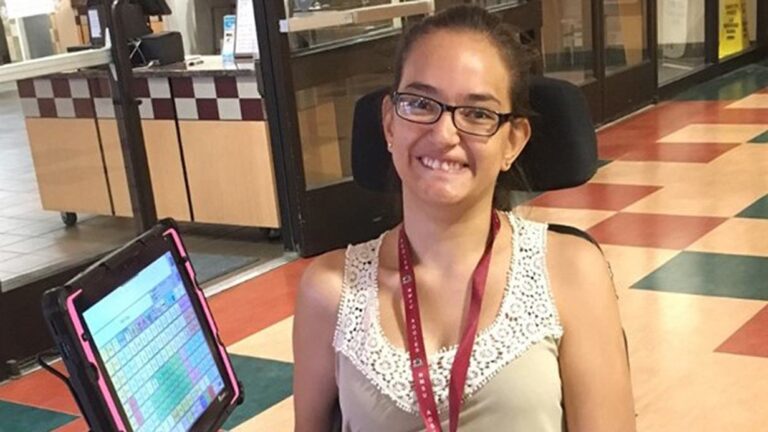 A young woman with glasses smiles while sitting in a wheelchair. She wears a sleeveless top with a lace collar and lanyard, and uses a communication device. The scene captures a moment from Arcys Story in a public building with tiled floors.