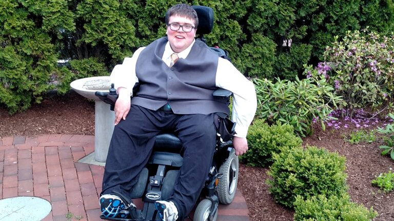 A young man in formal attire sits in a motorized wheelchair on a brick path in a garden, smiling at the camera. Surrounded by green shrubs and trees, he radiates positivity—an inspiring glimpse into Augusts Story.
