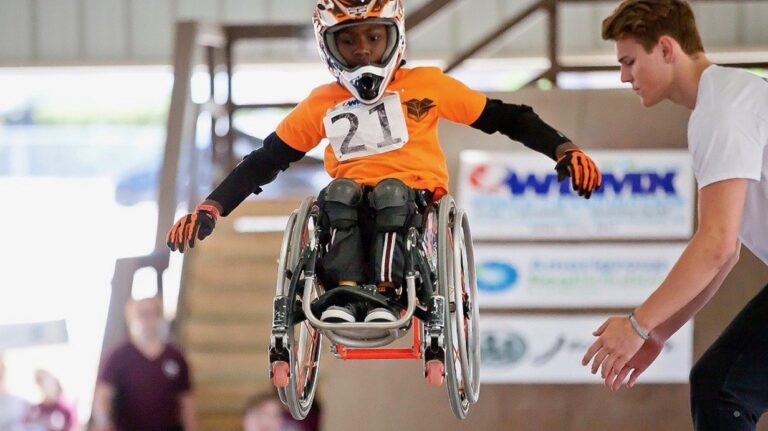 Young athlete Delmace, wearing a helmet and an orange shirt with the number 21, catches air while performing a jump at a skate park as an assistant reaches out nearby—an inspiring story of determination.