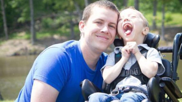 Garretts Story: A man in a blue shirt smiles and hugs a laughing child in a wheelchair outdoors, with green trees and water in the background.