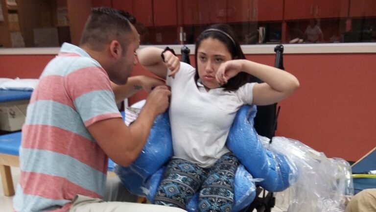 A man assists a young woman seated in a wheelchair with blue foam supports. As part of Genesiss Story, the woman looks focused and raises her arms while the man guides or instructs her.