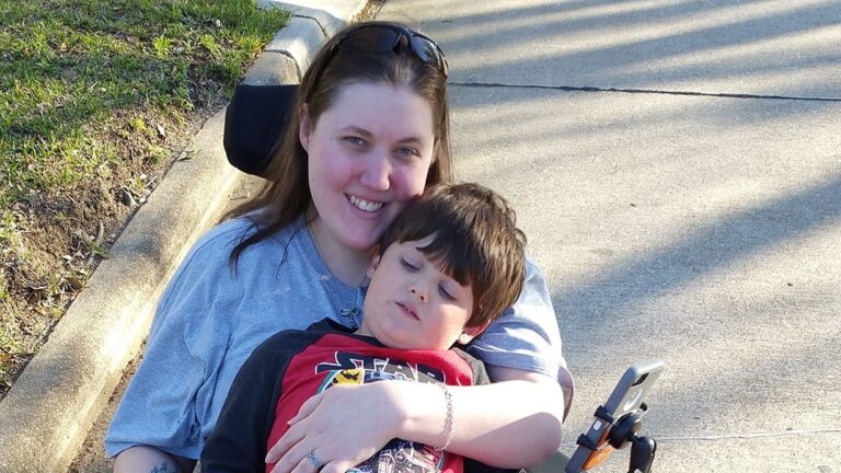 A woman in a wheelchair smiles while hugging a young boy in her lap. They are outdoors on a sunny day beside a curb, with green grass and shadows visible in the background—capturing a moment from Jessicas Story.