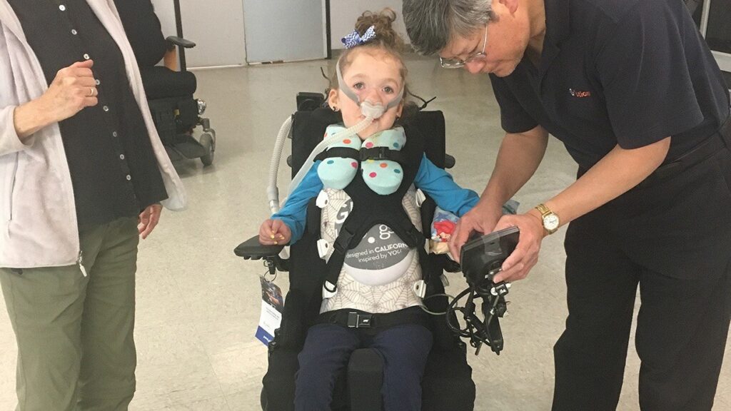 A young girl in a wheelchair, wearing a breathing mask and vest, is assisted by an older man holding a device. Another person stands nearby with their hand raised. The scene captures a moment from Lucys Story in an indoor setting.