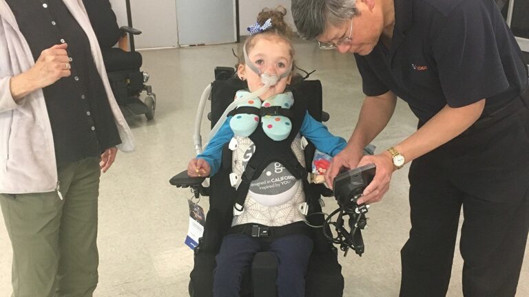 A young girl in a wheelchair, wearing a breathing mask and vest, is assisted by an older man holding a device. Another person stands nearby with their hand raised. The scene captures a moment from Lucys Story in an indoor setting.