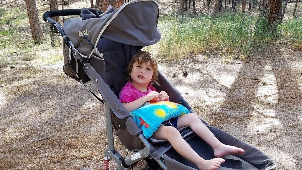 A young child sits in a stroller, holding a snack and a colorful blanket, surrounded by trees and dry ground in a forested area—this is part of Marleys Story.