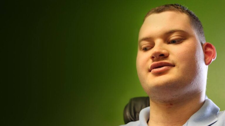 A young man with short hair and a light blue shirt sits in front of a green background, looking slightly downward with a neutral expression, capturing a moment from Michaels Story.