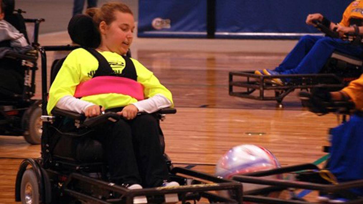 Sarahs Story: A young person in a wheelchair plays power soccer indoors, wearing a bright yellow shirt and guiding a large ball with a guard attached to the wheelchair. Other players in wheelchairs are also seen on the court.