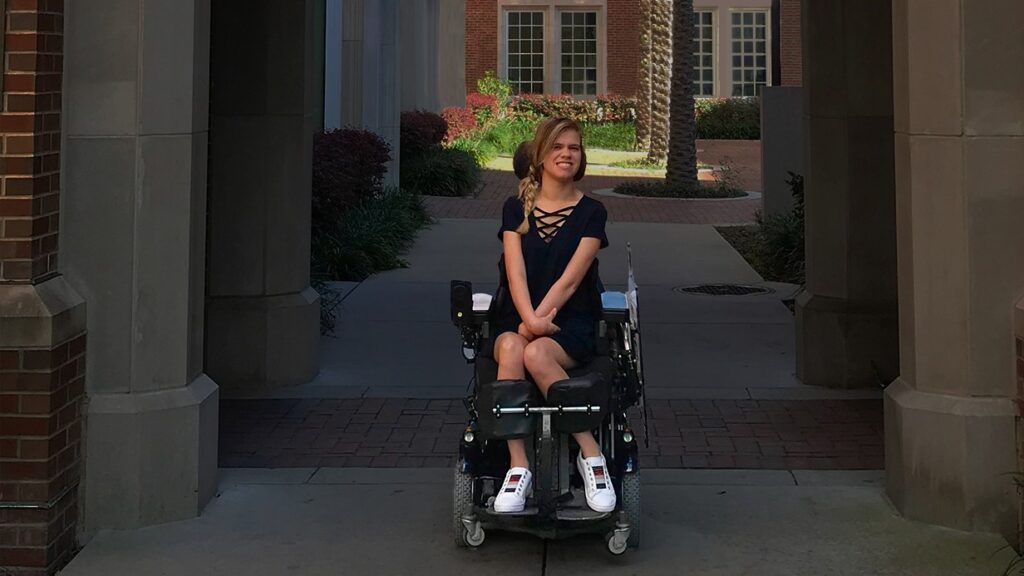 A young woman in a powered wheelchair sits under an archway outside, smiling and looking to the side. Sharing Delainas Story, she wears a navy dress and white sneakers as sunlight brightens the courtyard with plants and brick buildings behind her.