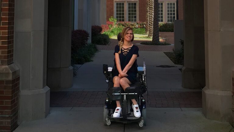 A young woman in a powered wheelchair sits under an archway outside, smiling and looking to the side. Sharing Delainas Story, she wears a navy dress and white sneakers as sunlight brightens the courtyard with plants and brick buildings behind her.