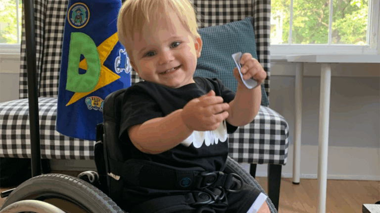 A smiling toddler with blonde hair sits in a wheelchair indoors, wearing a black shirt and shorts. Part of Donovans Story, the scene features a checkered chair with a blanket and a window revealing greenery outside.