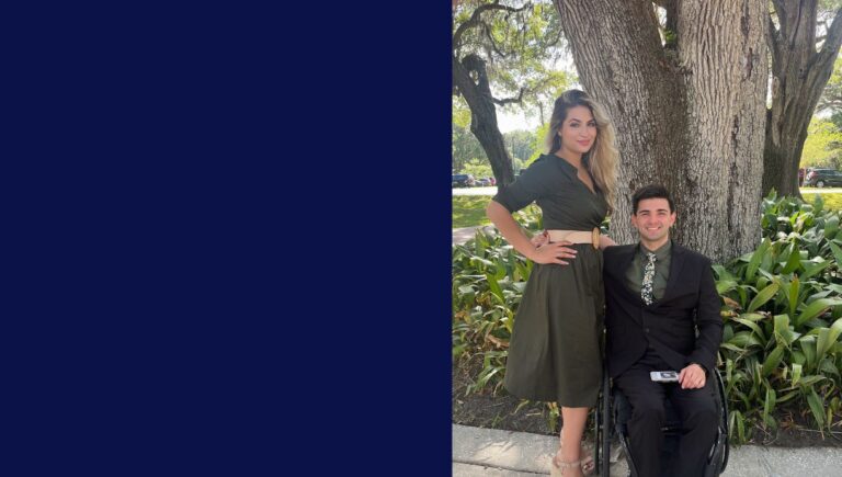 A woman stands next to a man in a wheelchair, both dressed formally in dark green outfits, posing outdoors near a large tree with greenery in the background, capturing a moment inspired by Dylans Story.