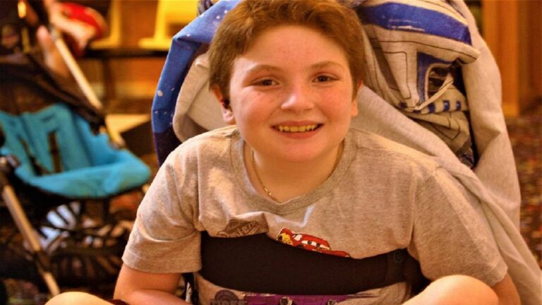 A smiling child with short brown hair sits in a wheelchair, wearing a light gray T-shirt. Indoors, colorful carpeting and a stroller can be seen in the background—part of Eldons Story.