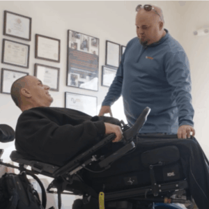 A NuMotion specialist speaks with a man reclining in a power wheelchair during a consultation. They are in an office with framed certificates on the wall, and both are engaged and smiling.