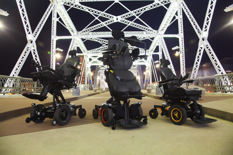 Three modern power recline wheelchairs are displayed side by side on a brightly lit steel bridge at night, with city lights visible in the background.