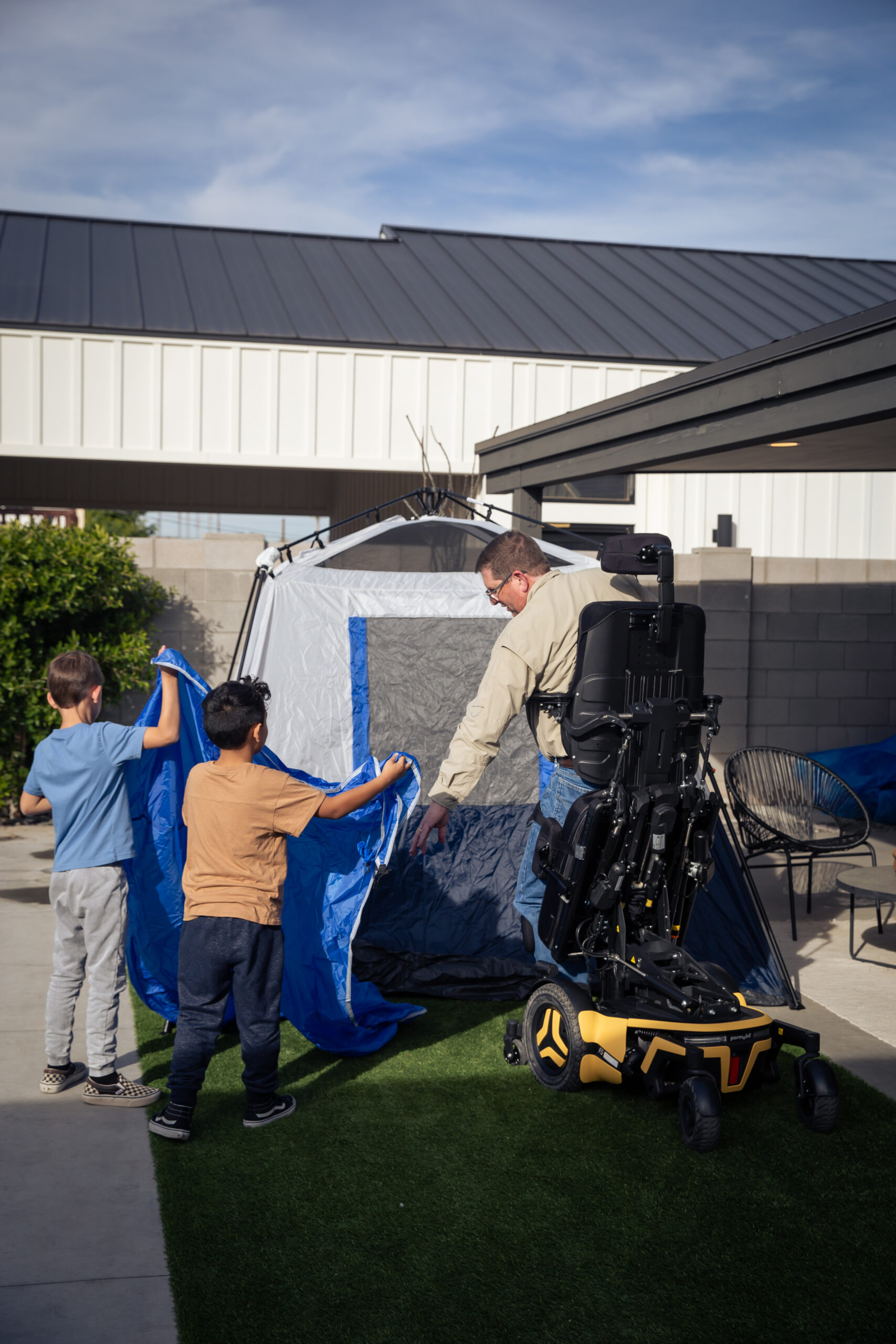 A man using a Complex Power Standing Wheelchair and two boys work together to set up a blue and gray tent on a grassy area next to a modern building under a clear sky.