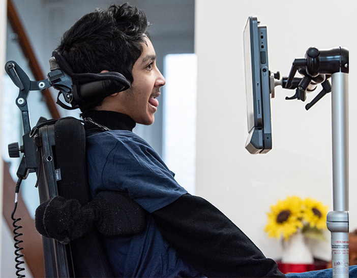 A young person in a power wheelchair uses a mounted speech-generating device. They are smiling and engaged with the screen, with a vase of sunflowers visible in the softly lit background.