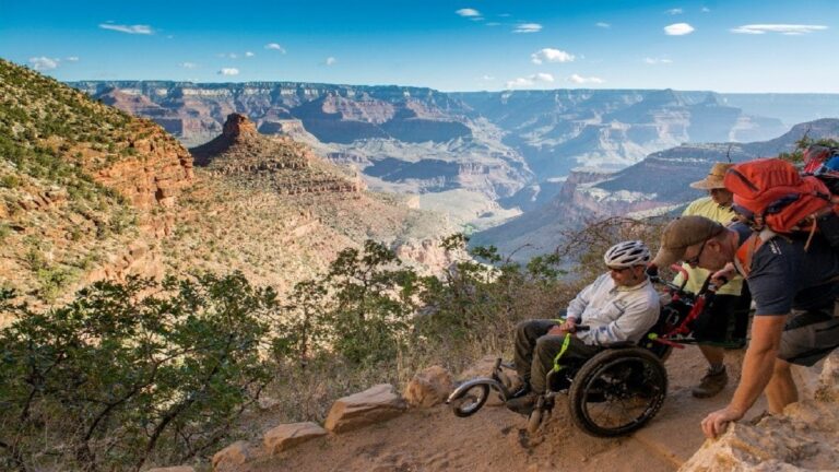 Geoffs Story: A person in a wheelchair, assisted by two people, navigates a rugged trail along the edge of the Grand Canyon, with expansive canyon views and blue sky in the background.