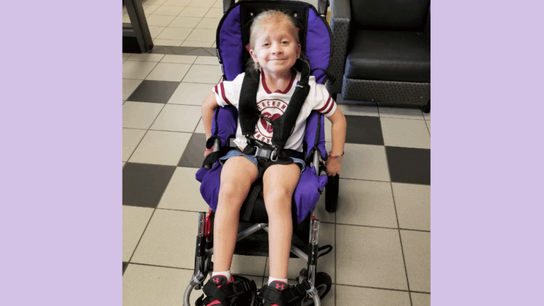A young girl with blonde hair smiles while sitting in a purple wheelchair. Sharing Haydens Story, she wears a white T-shirt with maroon trim, black shorts, and black shoes. The floor is tiled and gray chairs are in the background.