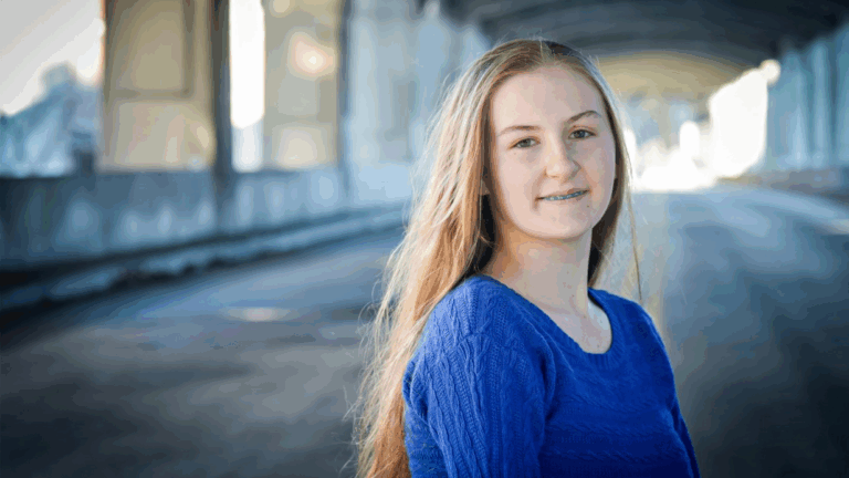 A young woman with long blonde hair and a blue sweater stands outdoors on an empty, sunlit street under an overpass, looking at the camera and smiling softly—capturing a moment from Heidis Story.
