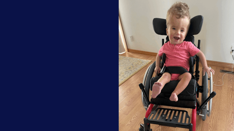 A young child with curly hair, wearing a pink shirt, smiles in a wheelchair on a wooden floor near a patterned rug and wall outlet. The left side features a solid navy blue background, highlighting Henrys Story.