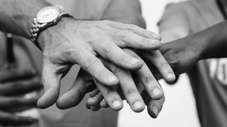 A close-up, black-and-white photo of several hands stacked on top of each other, symbolizing unity, hope, and teamwork. One person wears a watch.