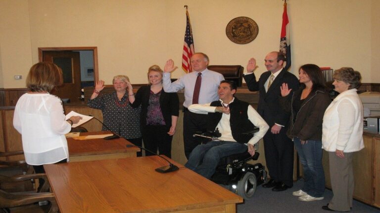 A group of seven people stands in a courtroom facing a woman holding papers, most with their right hands raised as if being sworn in. An American flag and a state flag are behind them. One man, central to Traviss Story, uses a wheelchair.