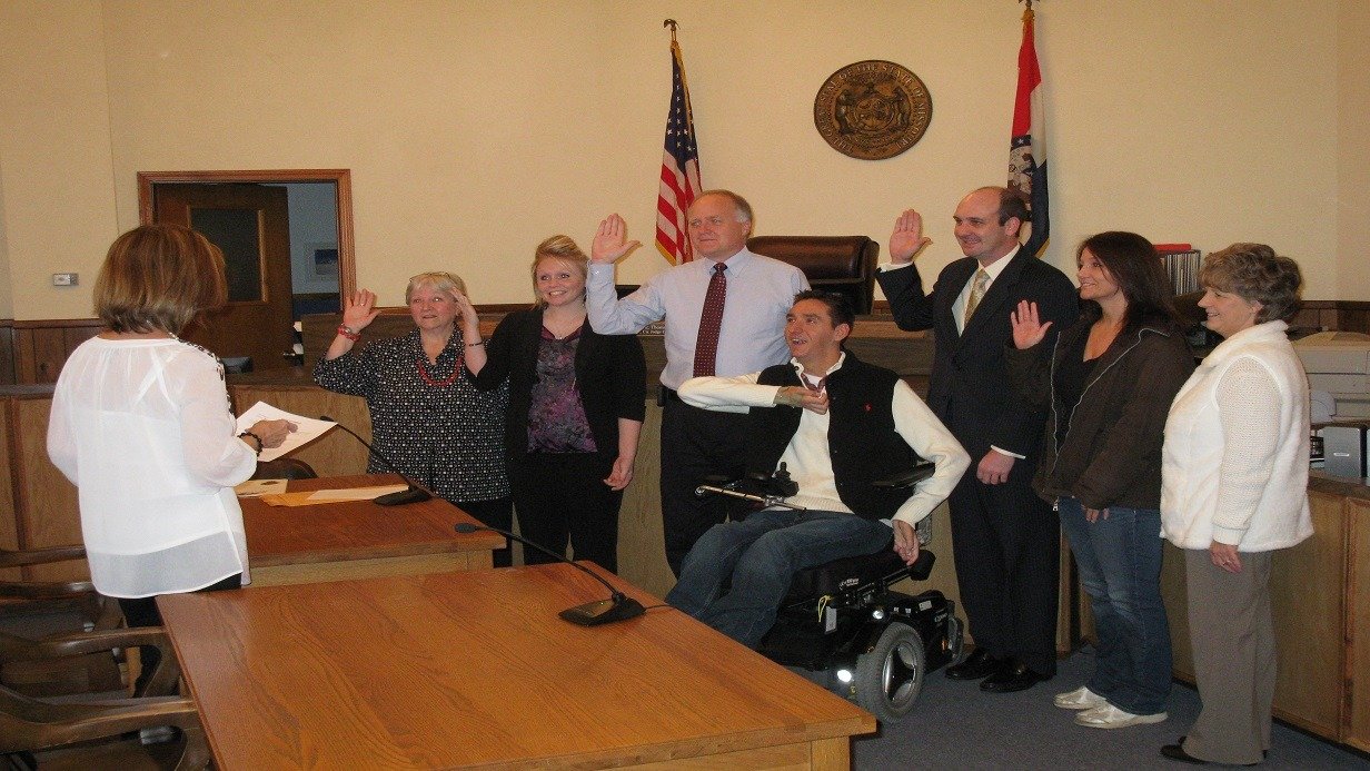 A group of seven people stands in a courtroom facing a woman holding papers, most with their right hands raised as if being sworn in. An American flag and a state flag are behind them. One man, central to Traviss Story, uses a wheelchair.