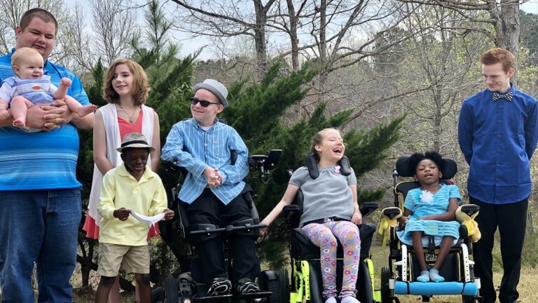 A group of seven children and teens, including some using wheelchairs, pose outside on a sunny day. They are smiling and dressed casually with trees and greenery in the background, sharing a joyful Evans Family story.