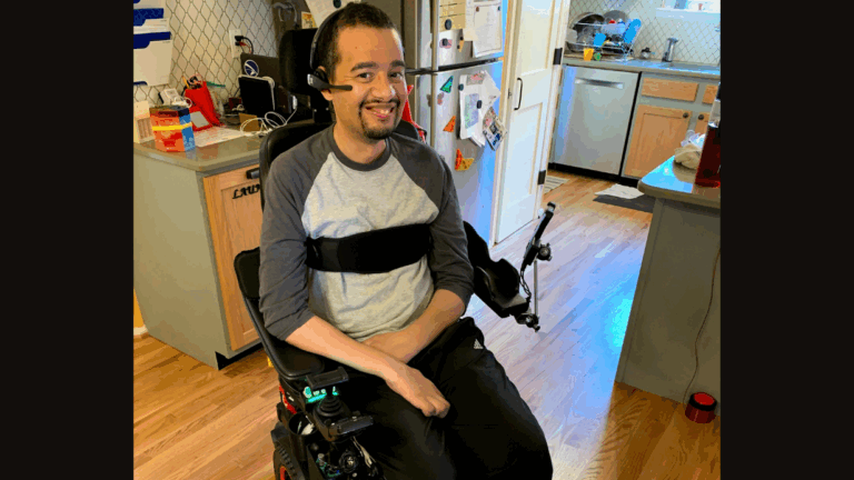 A smiling man sits in a motorized wheelchair in a bright kitchen with light wood floors, wearing a gray and black shirt. Jamess Story includes a device on his headrest and adaptive equipment on his chair.