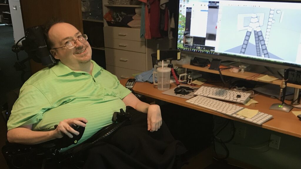 Jamie’s Story: A man in a green shirt sits at his desk, smiling at the camera. His workspace features a large monitor showing a 3D design, along with a keyboard, mouse, drink cup, and various papers.
