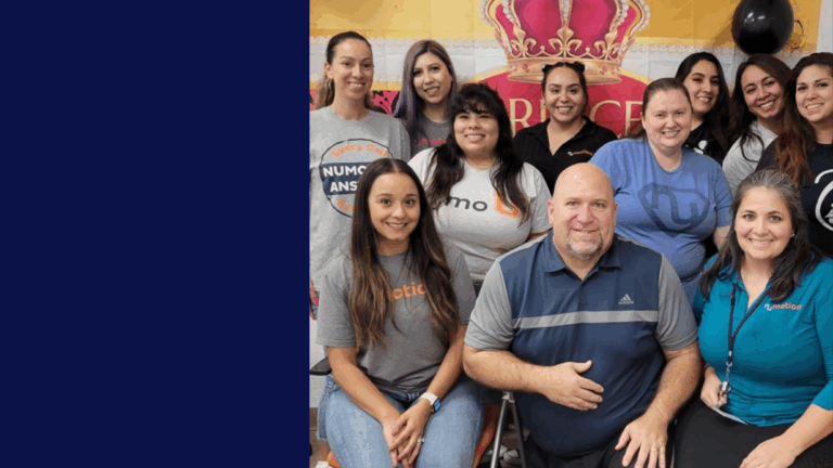 A group of thirteen smiling people, both men and women, pose together indoors in front of a decorative banner with a large pink crown. Most are casually dressed and gathered closely, sharing in Jeffs Story.