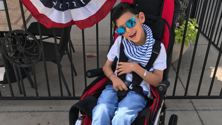 A smiling boy wearing sunglasses and a striped scarf sits in a red wheelchair outdoors near a black metal fence decorated with a red, white, and blue banner, sharing Jojos Story.