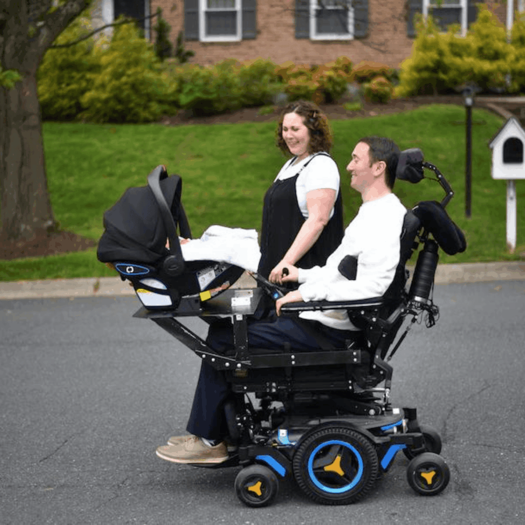 A man in a power wheelchair pushes a baby stroller attachment holding an infant car seat, accompanied by a smiling woman. They are outside on a residential street with trees and a brick house in the background.