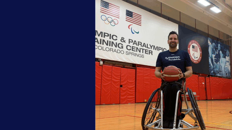 Josh’s Story: A man in a wheelchair holds a basketball on a gym court, smiling. Behind him are red mats, flags, and a large sign reading Olympic & Paralympic Training Center, Colorado Springs.
