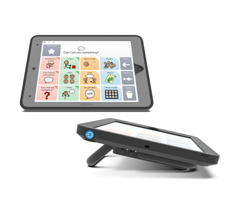 A Smartbox L-Series tablet with a stand displays a communication app featuring colorful symbol-based buttons for phrases like “I want,” “Please,” and “Help.” The screen helps users communicate needs and feelings.