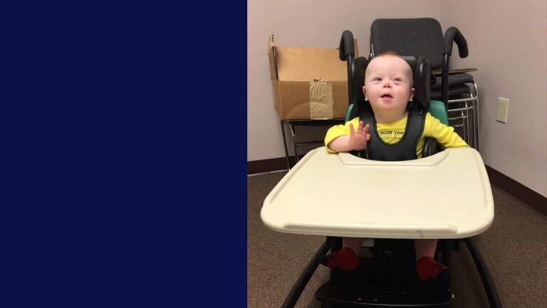 A young child with Down syndrome sits in a high chair, wearing a yellow Levis shirt and looking upward with a curious expression. Stacked chairs and a cardboard box in the background add detail to this heartfelt story.