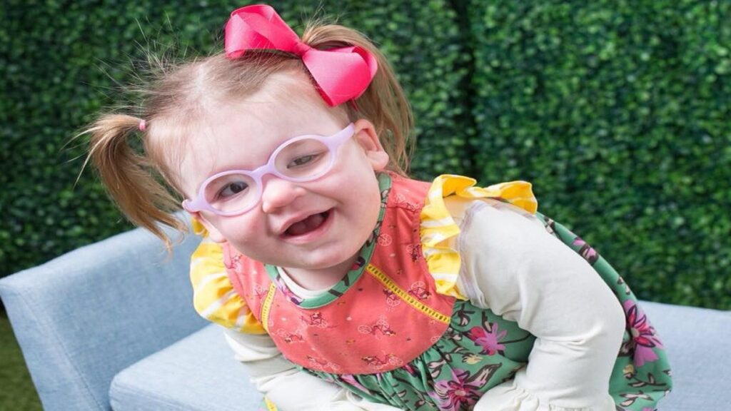 Londyns Story: A young girl with light hair in pigtails and a pink bow, wearing glasses and a colorful dress with ruffled sleeves, smiles as she leans forward on a light blue chair, set against a vibrant green leafy background.