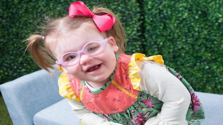 Londyns Story: A young girl with light hair in pigtails and a pink bow, wearing glasses and a colorful dress with ruffled sleeves, smiles as she leans forward on a light blue chair, set against a vibrant green leafy background.