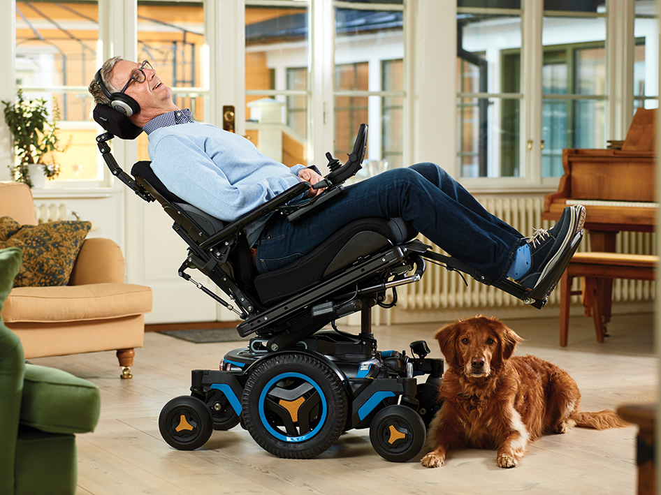 A smiling man reclines in a complex wheelchair with a joystick, inside a bright living room. A brown dog lies nearby on the floor. The room features large windows, cozy furnishings, and a piano.