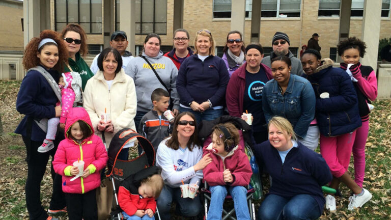 A diverse group of adults and children pose outside in front of a building, some smiling, some seated, including a child using a wheelchair. The ground is covered with leaves as everyone is dressed warmly—another moment from Hollies Story.